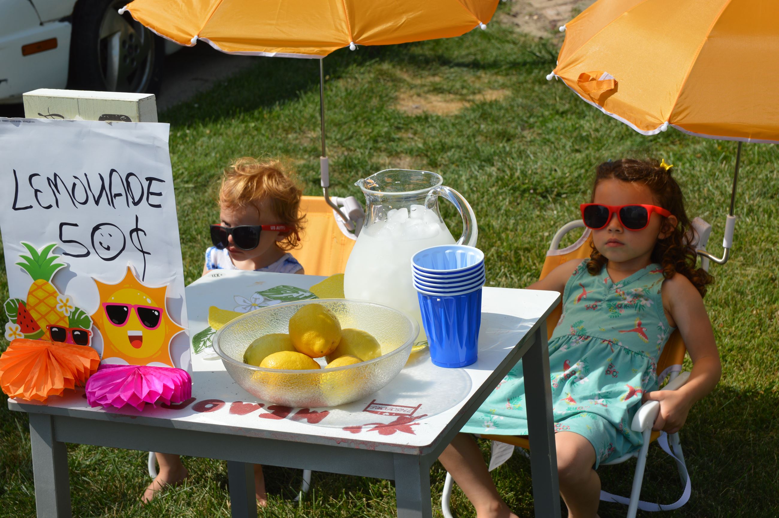 Two toddler girls recline in a lawn chair at their lemonade stand with their sunglasses on