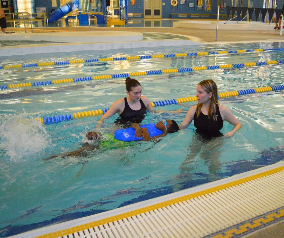 Two swim instructors help a little boy holding a blue boogie board learn how to paddle on his back.