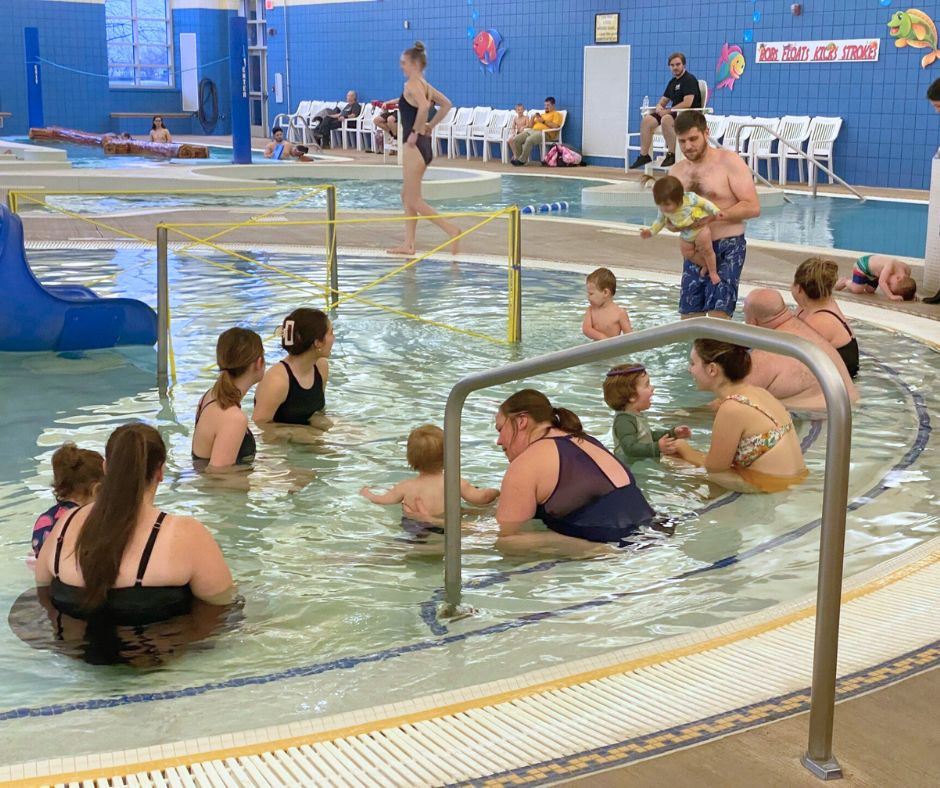 A group of parents with their toddlers sit in the kiddie pool before swim class begins. 