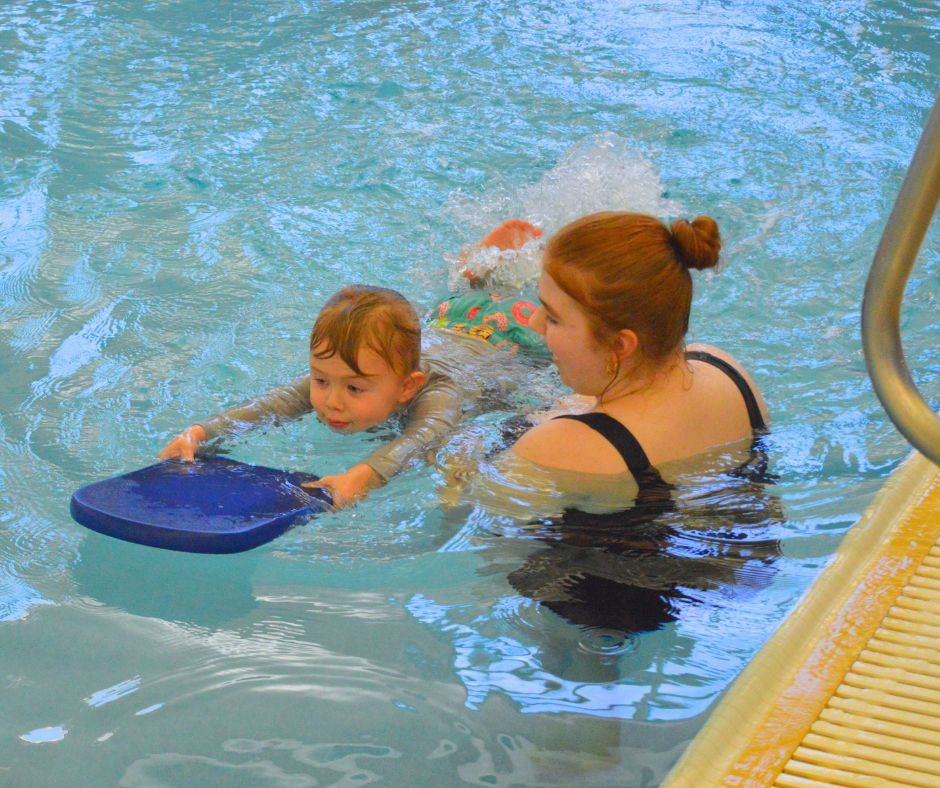 A swim instructor helps a little boy paddle with his feet while holding a blue boogie board. 