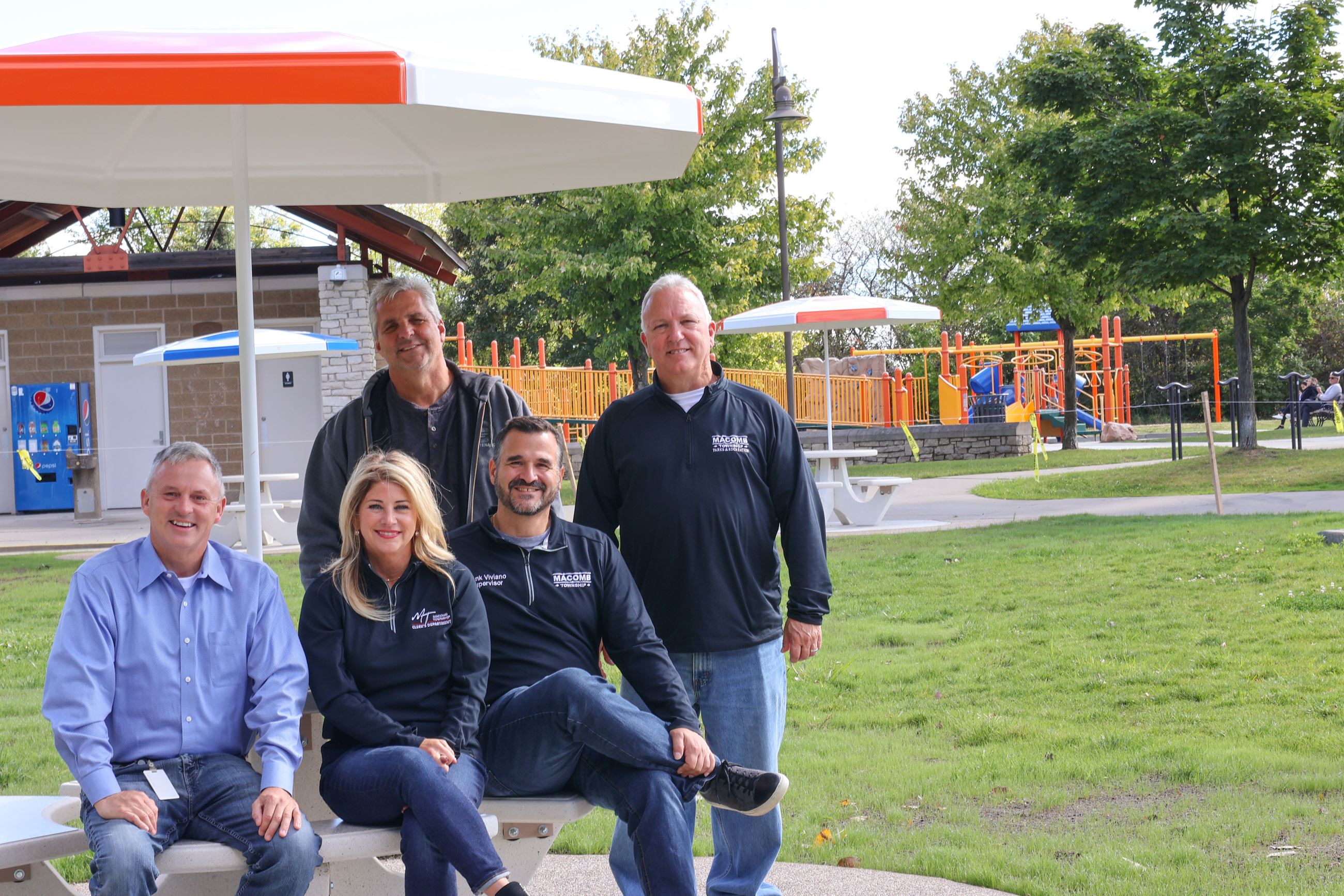 Five people sitting and smiling at a picnic table with an orange playscape behind them