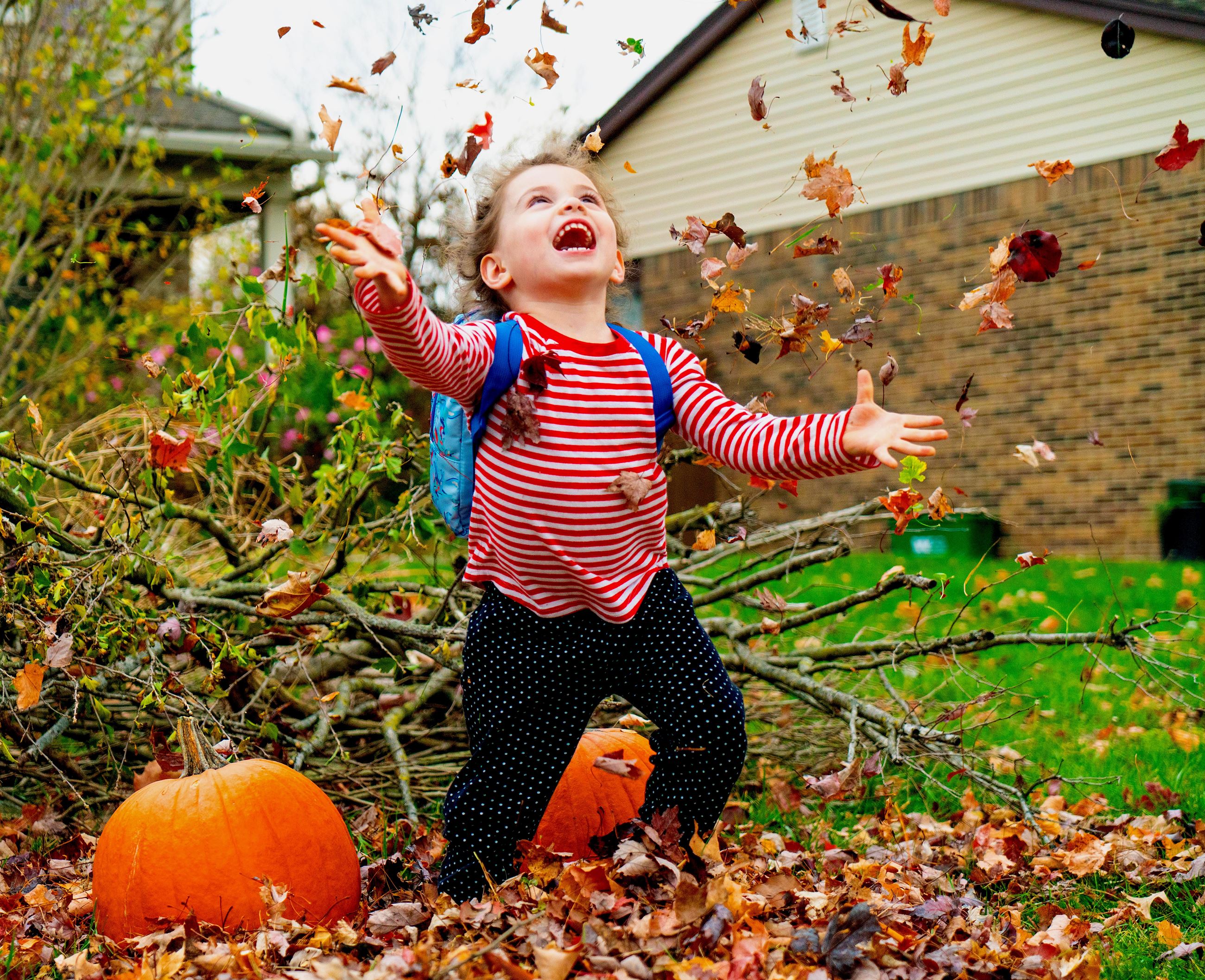 A little girl with a blue backpack stands in a pile of fallen leaves, throwing them up with a smile 
